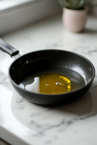A black saucepan on a white marble cooktop, with olive oil heating up over medium heat. The oil glistens, ready for the next step.