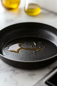A close-up shot of a black skillet on the white marble cooktop with olive oil heating. The surface glistens as it's ready for cooking the shrimp.