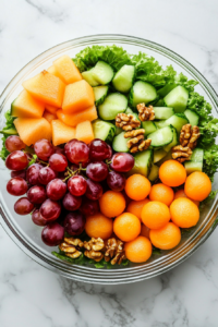 Close-up of a glass bowl on the white marble cooktop lined with fresh, crisp lettuce leaves. Vibrant melon balls, orange slices, halved grapes, and walnut halves are arranged in layers, creating a colorful and textured salad base.