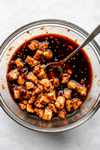Overhead shot of a glass mixing bowl with diced chicken pieces marinating in a dark, glossy mixture of soy sauce, chili vinegar, cornstarch, and ground ginger. A spoon stirs the ingredients evenly.