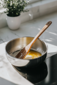 Close-up of a stainless steel saucepan on a white marble cooktop with melted butter bubbling gently. A wooden spatula rests on the edge, ready to stir.