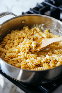 Close-up of a stainless steel saucepan on a white marble cooktop as freshly cooked semolina rotini is added to the cheesy sauce. A wooden spatula gently folds the pasta, coating it evenly.