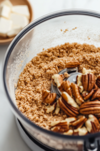A food processor on a wooden countertop, filled with graham crackers, pecans, light brown sugar, and salt. The crumbs are finely ground, ready for mixing with melted butter. Soft natural lighting highlights the texture of the ingredients.