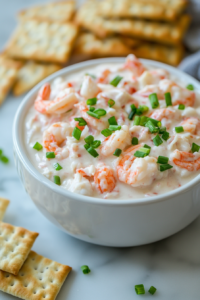 A close-up shot of a chilled serving bowl filled with creamy crab and shrimp dip on the white marble cooktop. The dip is garnished with extra chopped green onions and is surrounded by an assortment of crackers and toasted bread.