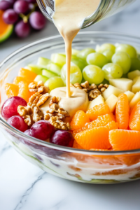 Close-up of the glass salad bowl on the white marble cooktop as the smooth, creamy dressing is being poured evenly over the layered melon, oranges, grapes, and walnuts. The salad glistens as it begins to marinate.
