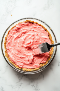 9-inch pie dish on a white marble countertop, as the velvety pink strawberry filling is poured over the chilled graham cracker crust and spread evenly with a spatula. The layers look smooth and inviting.
