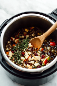 The black Instant Pot on a white marble cooktop is now filled with chicken broth, freshly chopped apple, a wild rice blend, dried cranberries, ground thyme, salt, and pepper. A wooden spoon stirs the colorful mixture before the chicken is added.