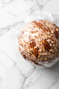 A close-up shot of the pecan-coated cheese ball wrapped in plastic wrap, resting on the white marble cooktop, ready to be chilled in the refrigerator for an hour.