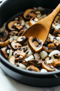 A black Instant Pot on a white marble cooktop set to sauté mode. Olive oil shimmers as sliced fresh mushrooms, diced onions, and minced garlic are sautéed. The vegetables soften, releasing a rich aroma while a wooden spoon stirs them gently.