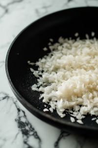 A black skillet on a white marble cooktop now filled with finely chopped white onions and minced garlic. The ingredients sizzle in the hot oil, turning golden and releasing a rich, fragrant aroma.