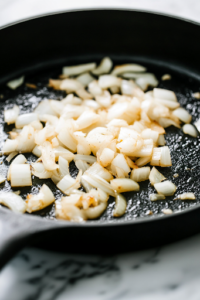 The black cast-iron skillet on the white marble cooktop, now with sliced onions and minced garlic being sautéed in olive oil. The mixture turns soft and fragrant, releasing a rich aroma.
