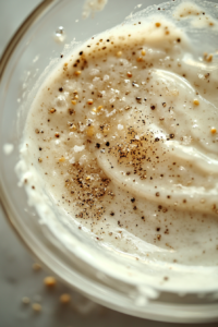 Close-up of a spoon sprinkling salt and white pepper into the creamy dressing in a glass bowl on the white marble cooktop. The seasonings dissolve, enhancing the flavor.
