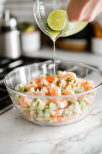 A close-up shot of a glass mixing bowl over the white marble cooktop with chopped shrimp inside. Freshly squeezed lime juice is being drizzled over the shrimp.