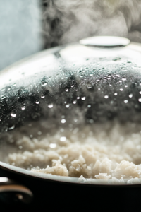 A glass lid covers the skillet as the dish simmers over low heat. Steam rises, and condensation forms on the lid while the rice slowly absorbs the liquid, ensuring a tender and flavorful result.