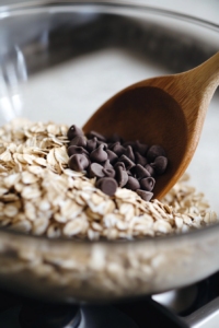 Close-up shot of rolled oats and semisweet chocolate chips being stirred into the thick cookie dough in the glass mixing bowl on the white marble cooktop. A wooden spoon is folding them in evenly.