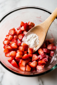 Diced strawberries and rhubarb are being tossed with flour in a glass mixing bowl on a white marble cooktop. A wooden spoon stirs gently to coat the fruit evenly.