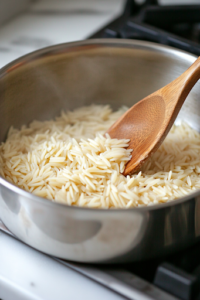 A stainless steel saucepan on the white marble cooktop, where orzo and jasmine rice are toasting in melted butter, turning golden brown. A wooden spoon stirs the mixture, ensuring even toasting.