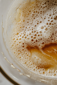 A glass mixing bowl on a white marble cooktop with sugar, oil, milk, egg, and vanilla being whisked into a smooth batter. The wet ingredients glisten under the kitchen light.