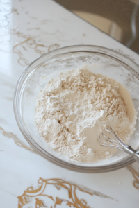 Close-up shot of a glass mixing bowl on the white marble cooktop, filled with all-purpose flour, baking powder, baking soda, and salt. A metal whisk is mid-motion, blending the dry ingredients.