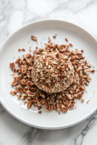 A close-up shot of a white plate over the white marble cooktop, filled with finely chopped pecans. The cheese ball is being gently rolled in the pecans, ensuring an even coating.