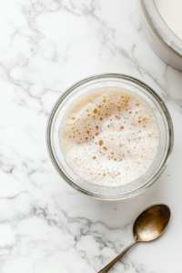 A close-up shot of a small glass bowl over the white marble cooktop, filled with warm water at 110°F. Active dry yeast is sprinkled on top, starting to dissolve and foam. A spoon rests nearby, ready for stirring.
