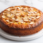 A top-down view of a golden apple almond cake freshly baked, topped with a sprinkle of sliced almonds and turbinado sugar. The cake sits on a white ceramic plate with parchment paper beneath it, all placed on a clean white marble countertop. The soft golden texture of the cake and the crispness of the topping shine in this simple and minimal scene, perfect for showcasing the dessert's lightness and flavor. The background enhances the cake's visual appeal, emphasizing its inviting look and texture.