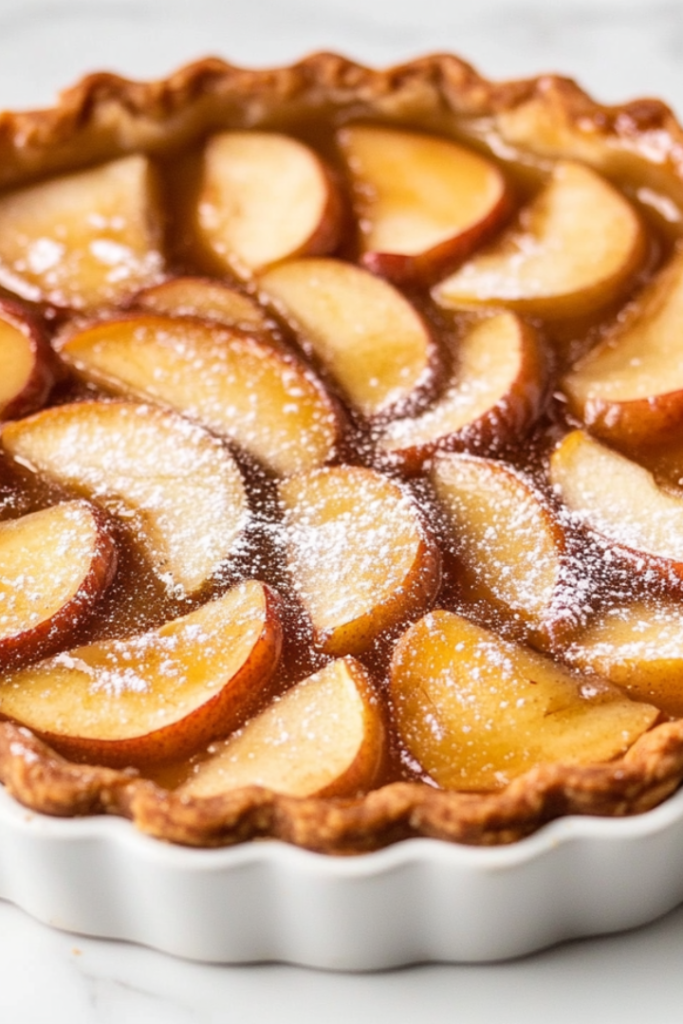 A fully baked apple custard tart beautifully presented in a white tart pan with removable bottom. The tart features golden-brown edges, perfectly layered apple slices, and a glossy glaze from apple jelly. Lightly dusted with confectioners’ sugar, the tart is placed on a white marble countertop, ready to serve. This image highlights the final baked product with a rustic yet elegant appearance, showcasing the results of the recipe in a clean and appetizing setup.