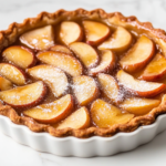 A fully baked apple custard tart beautifully presented in a white tart pan with removable bottom. The tart features golden-brown edges, perfectly layered apple slices, and a glossy glaze from apple jelly. Lightly dusted with confectioners’ sugar, the tart is placed on a white marble countertop, ready to serve. This image highlights the final baked product with a rustic yet elegant appearance, showcasing the results of the recipe in a clean and appetizing setup.