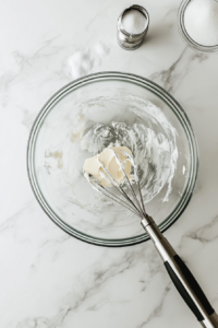 A top-down shot of a glass mixing bowl filled with softened unsalted butter and granulated sugar being beaten with a hand mixer. The mixture is creamy and fluffy, with a spatula and measuring cups on the marble countertop nearby. The scene is minimalistic, focusing on the smooth texture of the butter and sugar mixture as preparation for the cake.
