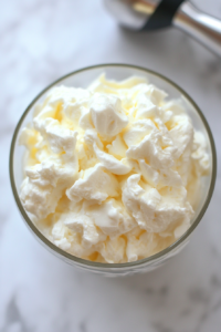 A clean glass bowl with whipped egg whites that have reached stiff peaks, with a hand mixer placed beside the bowl. The egg whites appear light and fluffy, forming a cloud-like texture. The white marble countertop enhances the clean aesthetic of the scene. The mixing process is captured with a focus on the airy texture of the egg whites, ready to be incorporated into the almond mixture for the cake.