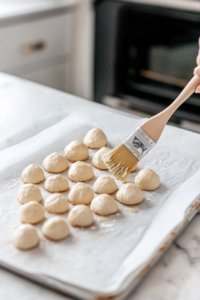 A top-down view of a hand using a pastry brush to coat the freshly baked golden brown bread bites with melted butter. The bread is then generously sprinkled with Italian seasoning, garlic powder, grated parmesan cheese, and finely chopped fresh parsley for added flavor. The Christmas tree-shaped bread arrangement sits on a white marble countertop, looking festive and flavorful, ready for serving.