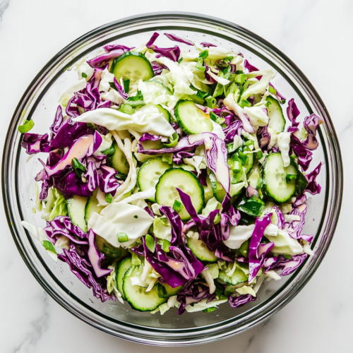 Top-down view of a colorful cabbage cucumber salad served in a clear glass bowl on a clean white marble countertop. The salad includes finely shredded white and red cabbage, thin cucumber slices, and chopped green onions tossed in a light sunflower oil and vinegar dressing. The dish looks fresh, crisp, and vibrant, ideal for any healthy meal.