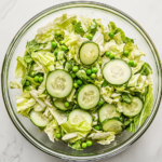 Top-down view of the complete cabbage cucumber sweet pea salad served in a large clear glass bowl on a white marble countertop. The vibrant green cabbage, crisp cucumber slices, sweet peas, and chopped green onions are evenly coated in a light vinegar and olive oil dressing, creating a fresh and colorful side dish perfect for warm days.