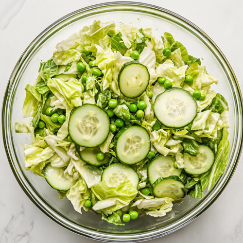 Top-down view of the complete cabbage cucumber sweet pea salad served in a large clear glass bowl on a white marble countertop. The vibrant green cabbage, crisp cucumber slices, sweet peas, and chopped green onions are evenly coated in a light vinegar and olive oil dressing, creating a fresh and colorful side dish perfect for warm days.