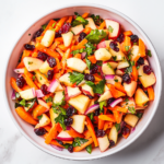 Top-down view of a fresh carrot apple salad served in a white bowl on a white marble countertop. The salad includes coarsely grated carrots, diced red apples, dried cranberries, and finely chopped purple onion all tossed in a creamy mayo dressing. This colorful, crunchy salad is simple, vibrant, and full of texture and flavor.