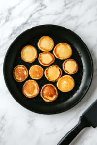 A black non-stick skillet on a white marble countertop has small, round pancakes cooking to golden perfection. A stainless steel spatula is lifting one pancake, revealing its evenly browned surface. The skillet has a light coating of oil, ensuring a crispy yet fluffy texture, ideal for making breakfast pancake sandwiches.