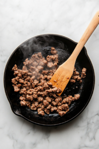 A black skillet sits on a white marble countertop with Italian sausage sizzling over medium heat. The sausage is broken into small crumbles and browned evenly, releasing flavorful oils. A wooden spatula rests in the skillet, ready to stir the meat, ensuring it cooks thoroughly before mixing with other ingredients.