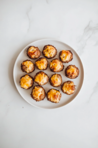 A plate filled with golden brown, freshly baked sausage cheese balls arranged on a white ceramic plate. The crispy, slightly cracked surface reveals melted cheddar cheese and flavorful sage sausage inside. The plate is placed on a white marble countertop with a minimal, clean background, making the delicious appetizer the main focus.