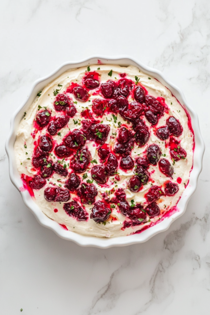 Top-down view of the finished cranberry jalapeño cream cheese dip presented on a white ceramic dish over a white marble countertop. The deep red cranberry topping with visible jalapeño specks is layered over creamy white cream cheese. It’s beautifully chilled and served with crispy crackers, making it a perfect party appetizer with bold flavors.