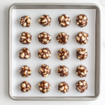 Top-down view of finished chocolate peanut clusters arranged neatly on a sheet of parchment paper placed over a baking tray. The glossy chocolate covers each peanut-filled cluster, giving them a rich and indulgent appearance. These homemade treats are cooling and ready to enjoy, showcasing the perfect blend of white and semisweet chocolate with roasted peanuts.