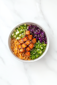 A top-down shot of a bowl of finished ramen, topped with crispy tofu, sautéed vegetables, sesame seeds, and chopped scallions, all served on a white marble countertop. The vibrant colors of the toppings contrast beautifully with the noodles and sauce, showcasing the dish in a simple yet inviting way. The clean setup draws attention to the completed, flavorful dish.