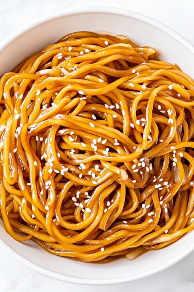 A delicious bowl of stir-fried soy garlic noodles served in a clean white ceramic bowl placed on a white marble countertop. The noodles are golden brown and glossy, coated in a savory soy and teriyaki sauce, and garnished with white sesame seeds. Bits of sautéed garlic, ginger, and onions are visible, making it look rich and flavorful. The minimal, bright background keeps the focus on the warm and comforting dish, ready to be served hot.
