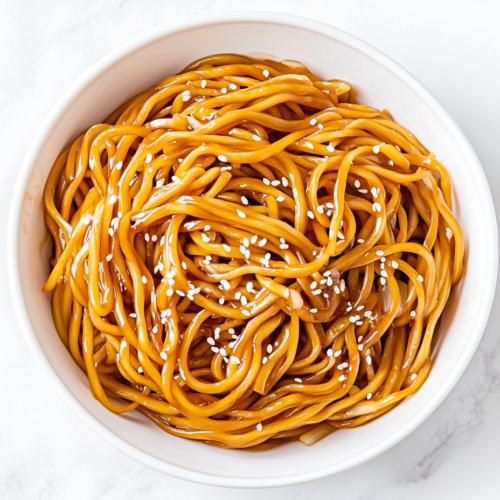 A delicious bowl of stir-fried soy garlic noodles served in a clean white ceramic bowl placed on a white marble countertop. The noodles are golden brown and glossy, coated in a savory soy and teriyaki sauce, and garnished with white sesame seeds. Bits of sautéed garlic, ginger, and onions are visible, making it look rich and flavorful. The minimal, bright background keeps the focus on the warm and comforting dish, ready to be served hot.