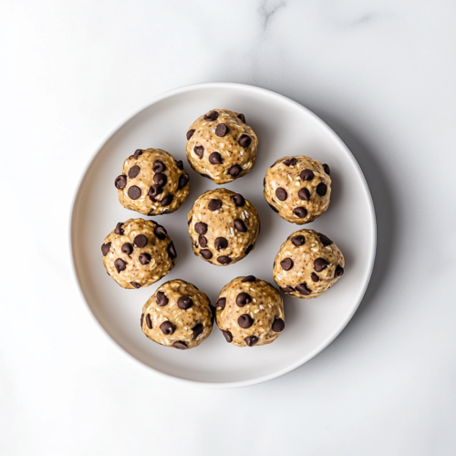 A top-down view of finished no-bake energy bites made with creamy cashew butter, oats, flaxseed, and mini chocolate chips. The bites are perfectly round and arranged on a white ceramic plate, highlighting their chewy texture and speckled chocolate chips. The clean, white marble background enhances the wholesome and homemade look of this protein-rich snack.