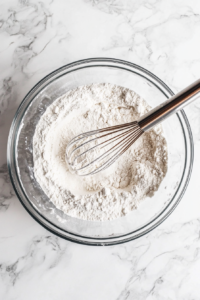 Top-down view of a mixing bowl with a whisk stirring in all-purpose flour, baking powder, sea salt, and milk into the cake batter. A small measuring cup of milk rests on the countertop next to the bowl, while the dry ingredients are scattered around. The marble countertop provides a clean, simple backdrop, focusing on the smooth texture being achieved in the batter.