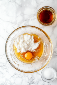 A top-down shot of a mixing bowl filled with almond flour, cornstarch, baking powder, and salt, ready to be combined with the egg yolk mixture. A whisk rests next to the bowl, and a measuring spoon with salt sits nearby. The white marble countertop provides a simple backdrop, allowing the ingredients to stand out. The scene captures the crucial step of mixing the dry ingredients into the egg yolk mixture for a smooth cake batter.