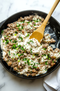 A black skillet on a white marble countertop containing browned Italian sausage mixed with soft, melting cream cheese, minced garlic, chopped green onions, and fresh parsley. A wooden spatula is stirring the mixture, making it creamy and well-blended, ready for stuffing into the mushrooms.