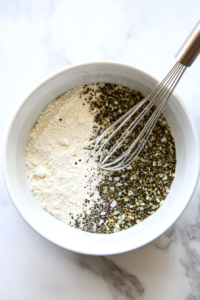 A large white mixing bowl on a white marble countertop contains a mixture of all-purpose flour, salt, black pepper, onion powder, and baking powder. A metal whisk rests inside, showing that the ingredients are being evenly combined. The scene is minimal, keeping the focus on the essential dry ingredients.