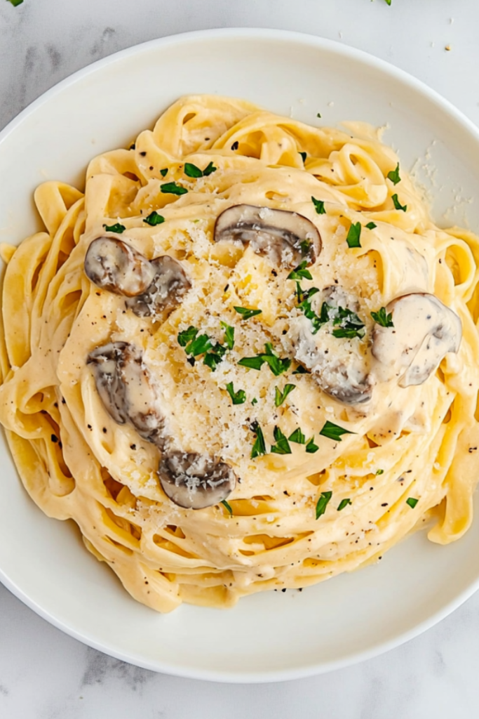 Top-down view of creamy mushroom fettuccine pasta served on a white ceramic plate over a white marble countertop. The pasta is generously coated in a rich cream sauce filled with mushrooms and parmesan, then garnished with fresh chopped parsley. The dish looks elegant, warm, and inviting, perfect for a cozy dinner.