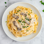 Top-down view of creamy mushroom fettuccine pasta served on a white ceramic plate over a white marble countertop. The pasta is generously coated in a rich cream sauce filled with mushrooms and parmesan, then garnished with fresh chopped parsley. The dish looks elegant, warm, and inviting, perfect for a cozy dinner.